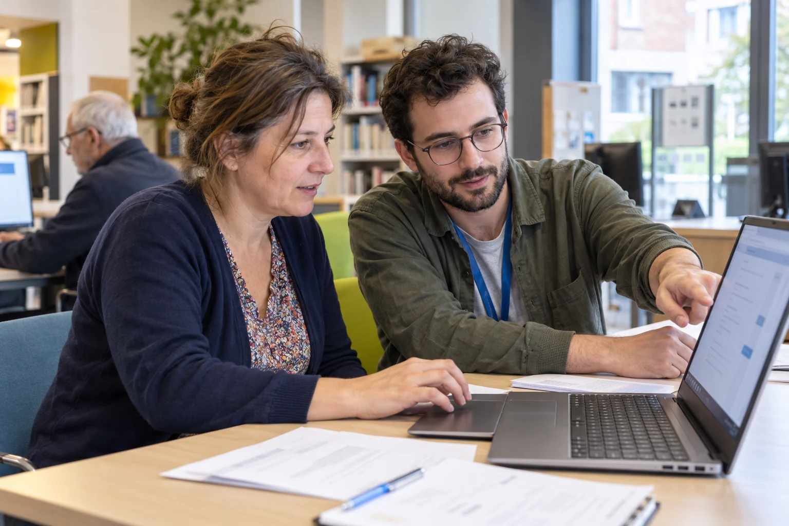 Une femme utilise un ordinateur portable avec l’aide d’un accompagnant dans un espace public numérique.